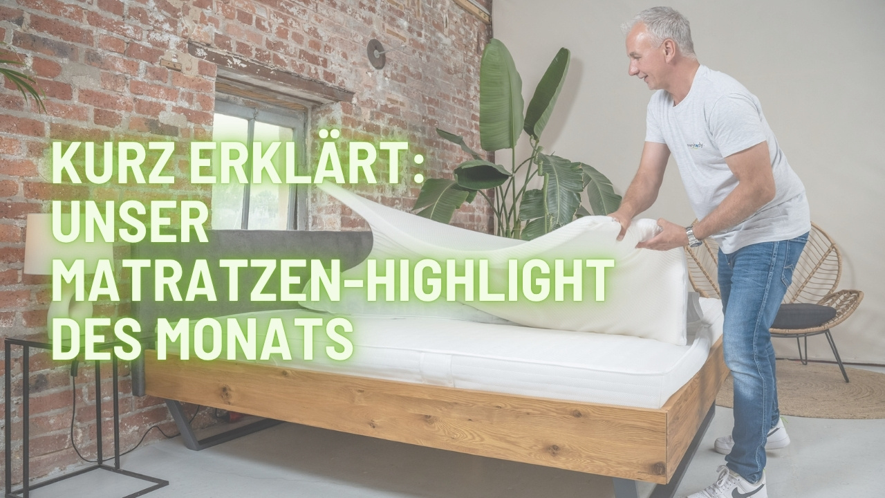 Heiko steht vor Bettgestell und hat einen Topper in der Hand. Auf dem Bild ist Schrift "die Matratze des Monats"